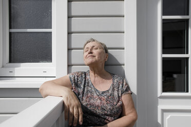 Entspannte ältere Frau sitzt auf der Veranda im Sonnenschein - KMKF00917
