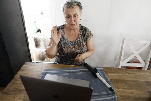 Senior woman sitting at table at home with laptop and curling iron - KMKF00923