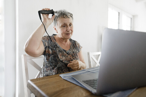 Ältere Frau, die zu Hause am Tisch sitzt, einen Laptop benutzt und sich die Haare macht, lizenzfreies Stockfoto