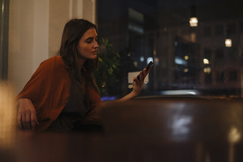 Junge Frau sitzt am Tisch in einem Restaurant und schaut auf ihr Handy - KNSF05759