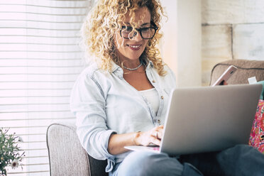 Smiling woman using laptop and cell phone on couch at home - SIPF01946