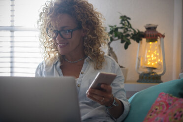 Smiling woman using laptop and cell phone on couch at home - SIPF01947