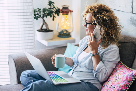 Frau auf der Couch zu Hause mit Kaffeetasse, Laptop und Mobiltelefon, lizenzfreies Stockfoto