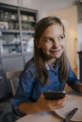 Smiling girl at home sitting at table with cell phone - KNSF05826