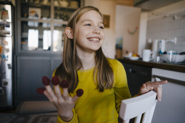 Glückliches Mädchen sitzt am Küchentisch zu Hause und spielt mit Himbeeren - KNSF05857