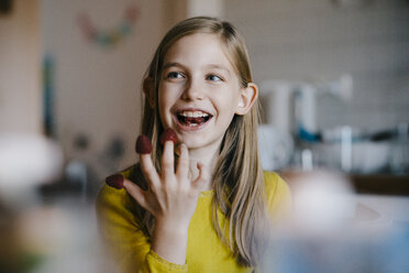 Happy girl sitting at kitchen table at home playing with raspberries - KNSF05859