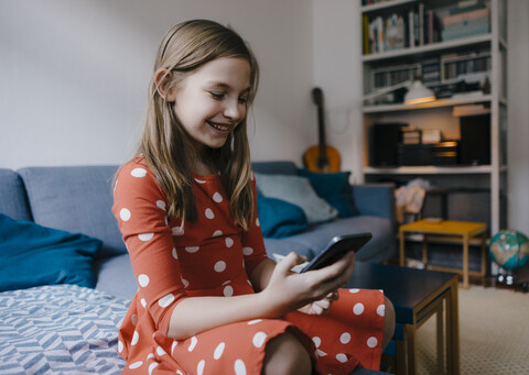 Glückliches Mädchen sitzt zu Hause auf der Couch und benutzt ein Mobiltelefon, lizenzfreies Stockfoto