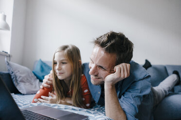 Happy father and daughter lying on couch at home using laptop - KNSF05885