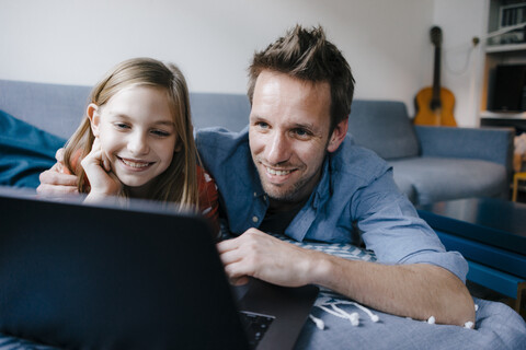 Glücklicher Vater und Tochter liegen auf der Couch zu Hause und benutzen einen Laptop, lizenzfreies Stockfoto