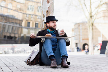 Male skateboarder sitting in city square with takeaway drink, Freiburg, Baden-Wurttemberg, Germany - CUF50836