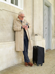 Senior man with wheeled suitcase leaning against building looking at his watch, Copenhagen, Hovedstaden, Denmark - CUF50908