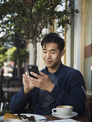 Lächelnder chinesischer Mann, der in einem Café im Freien eine SMS auf seinem Handy schreibt - BLEF03066