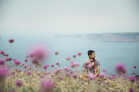 Kaukasischer Mann sitzt in einem Blumenfeld in der Nähe des Ozeans, lizenzfreies Stockfoto