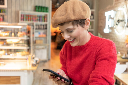 Mittlere erwachsene Frau mit Baskenmütze, die im Café auf ihr Smartphone schaut - CUF51265