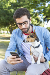 Porträt eines jungen Mannes, der mit seinem Hund auf einer Parkbank sitzt und auf sein Smartphone schaut - WPEF01504