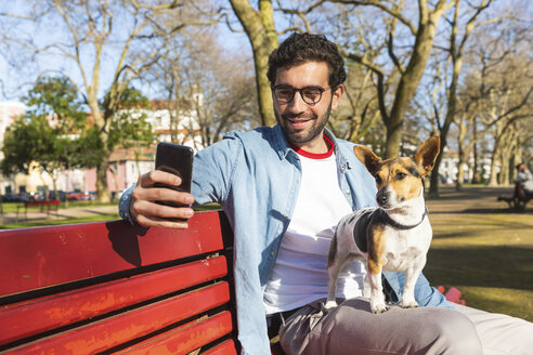 Porträt eines jungen Mannes, der mit seinem Hund auf einer Parkbank sitzt und ein Selfie mit seinem Smartphone macht - WPEF01514