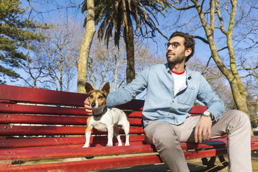 Zufriedener junger Mann sitzt auf einer Parkbank und streichelt seinen Hund - WPEF01516