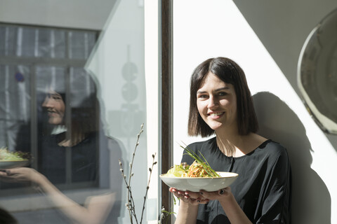 Lächelnde junge Frau, die bei Sonnenschein am Fenster steht und einen Salatteller hält, lizenzfreies Stockfoto