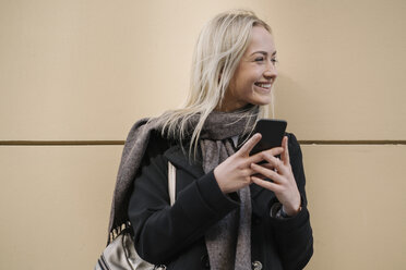 Smiling young woman with cell phone at a wall - AHSF00364