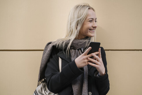 Lächelnde junge Frau mit Mobiltelefon an einer Wand - AHSF00364