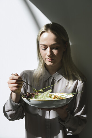 Junge Frau steht an einer Mauer in der Sonne und isst einen Salat, lizenzfreies Stockfoto