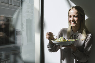 Smiling young woman standing at the window in sunshine eating a salad - AHSF00382