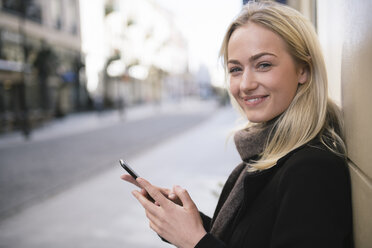 Porträt einer lächelnden jungen Frau mit Mobiltelefon in der Stadt - AHSF00389