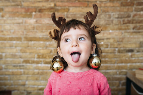 Porträt eines kleinen Mädchens, das die Zunge herausstreckt und ein Stirnband mit Rentiergeweih und Weihnachtskugeln trägt, lizenzfreies Stockfoto