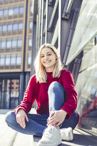 Porträt einer lächelnden jungen Frau, die im Sonnenlicht an der Wand sitzt, lizenzfreies Stockfoto