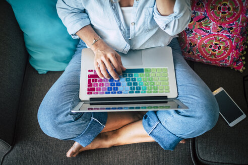 Woman using laptop with multicoloured keyboard on couch at home - SIPF01961