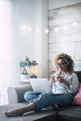 Woman on couch at home with coffee mug and laptop - SIPF01966