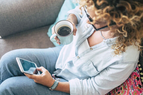 Frau auf der Couch zu Hause mit Kaffeebecher und Mobiltelefon, lizenzfreies Stockfoto