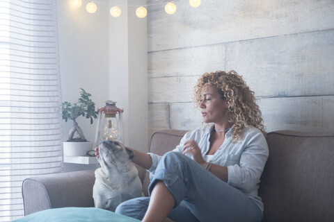 Frau sitzt zu Hause auf der Couch und streichelt einen Mops, lizenzfreies Stockfoto