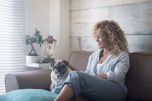 Woman sitting on couch at home petting pug dog - SIPF01974