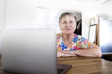 Porträt einer lächelnden älteren Frau, die zu Hause am Tisch sitzt und einen Laptop benutzt - KMKF00963