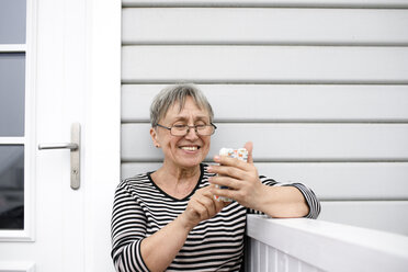 Glückliche ältere Frau, die auf der Veranda ihres Hauses ein Mobiltelefon benutzt - KMKF00965