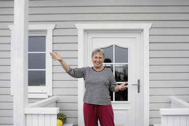 Glückliche ältere Frau steht auf der Veranda ihres Hauses - KMKF00967