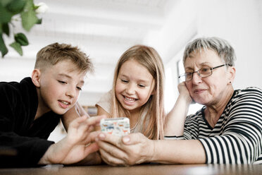 Glückliche Großmutter und Enkelkinder, die zu Hause ein Mobiltelefon benutzen - KMKF00972