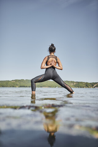 Junge Frau beim Yoga, stehend am Seeufer, Kriegerpose, Rückansicht, lizenzfreies Stockfoto