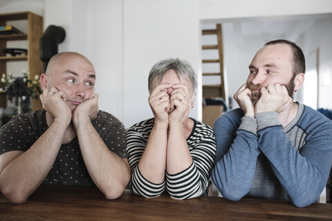 Porträt erwachsener Söhne mit älterer Mutter, die zu Hause am Tisch sitzen und den Kopf in den Händen halten, lizenzfreies Stockfoto