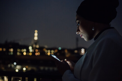 Junge Geschäftsfrau schaut nachts auf dem Balkon ihres Büros auf ihr Smartphone, lizenzfreies Stockfoto