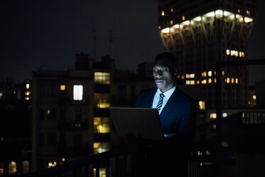 Junger Geschäftsmann mit Laptop auf dem nächtlichen Balkon seines Büros - CUF51336