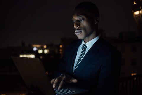 Junger Geschäftsmann tippt nachts auf dem Balkon seines Büros auf einem Laptop, Nahaufnahme, lizenzfreies Stockfoto