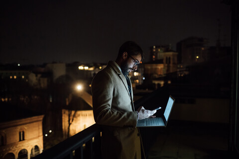 Mittlerer erwachsener Geschäftsmann, der nachts auf dem Balkon seines Büros auf sein Smartphone schaut, lizenzfreies Stockfoto