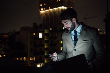 Mid adult businessman looking at smartphone on office balcony at night - CUF51343