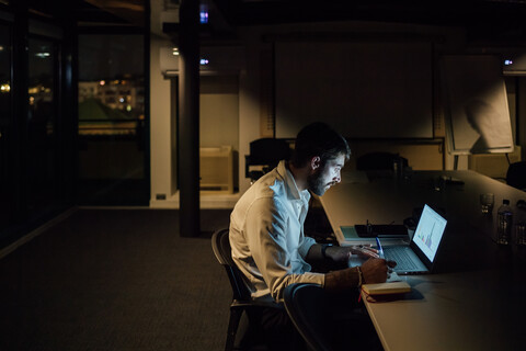 Mittlerer Erwachsener Geschäftsmann im Büro bei Nacht mit Blick auf den Laptop, lizenzfreies Stockfoto