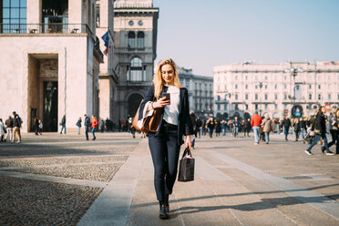 Young female tourist with shopping bags strolling and looking at smartphone in city square, Milan, Italy - CUF51376