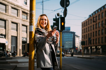 Young woman with smartphone laughing at tram station, Milan, Italy - CUF51383