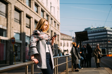 Junge Frau schaut an der Straßenbahnhaltestelle auf ihr Smartphone - CUF51384