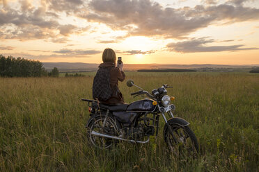 Kaukasische Frau mit Motorrad im Feld beim Fotografieren des Sonnenuntergangs - BLEF03251